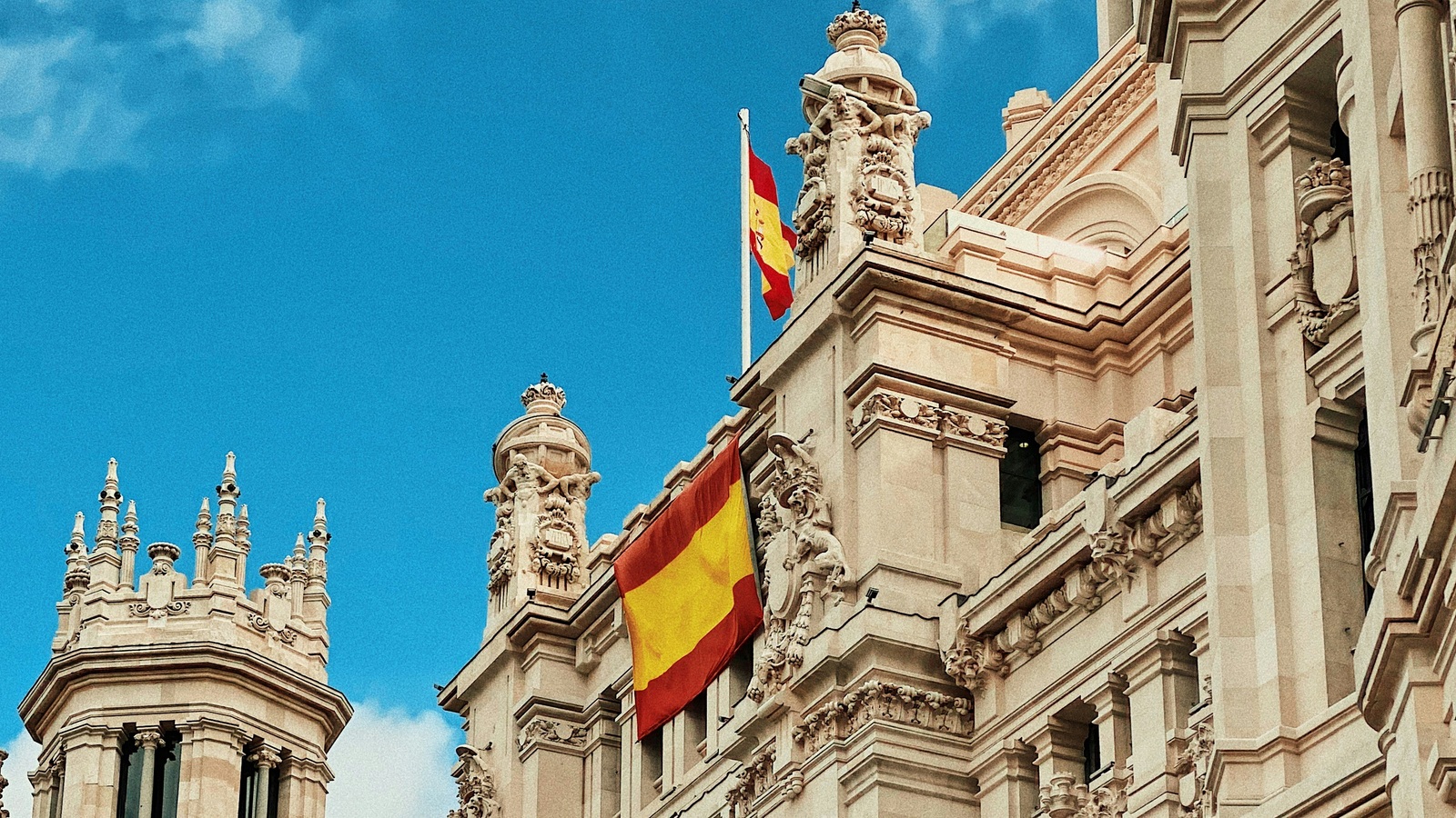 The façade of the Palacio de Cibeles in Madrid, with two Spanish flags and ornate turrets against a blue sky