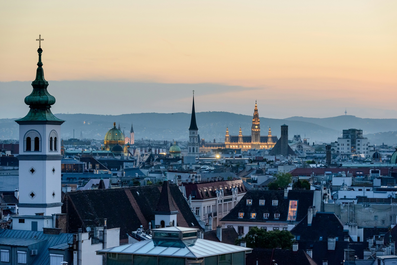 I tetti di Vienna al tramonto: in primo piano il campanile verde di una chiesa, sullo sfondo il Municipio illuminato e le colline
